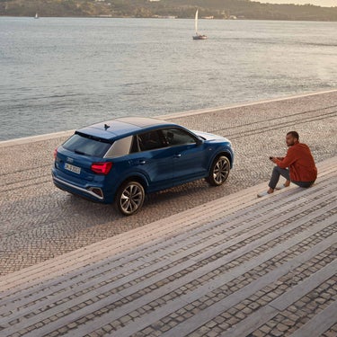 Ein blauer Audi Q2 schräg von hinten an einer Uferpromenade, Mann in oranger Jacke sitzt auf den Stufen und guckt zur Seite.
