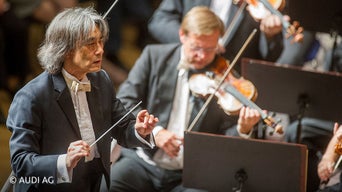DirigentKent Nagano beim Dirigieren. Im Hintergrund ist der Geiger des Orchesters zu sehen.