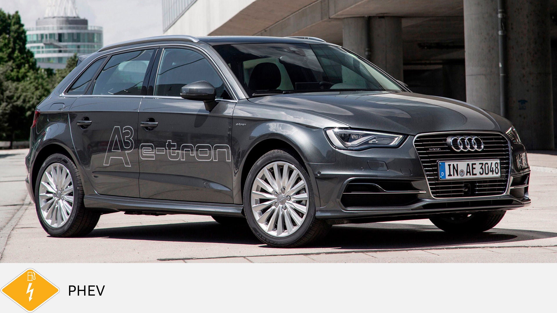 Grauer Audi A3 e-tron steht auf Betonfläche, e-tron-Schriftzug an der Seite, unten gelbes PHEV-Symbol mit Zapfsäule und Blitz.
