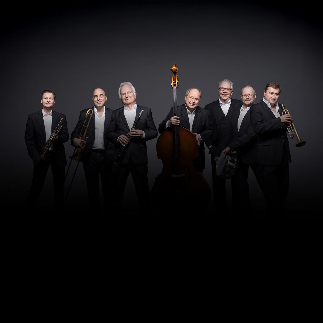 Sieben Musiker in schwarzen Anzügen stehen mit Trompete, Posaune, Klarinette, Kontrabass und Schlagzeug vor grauem Hintergrund.