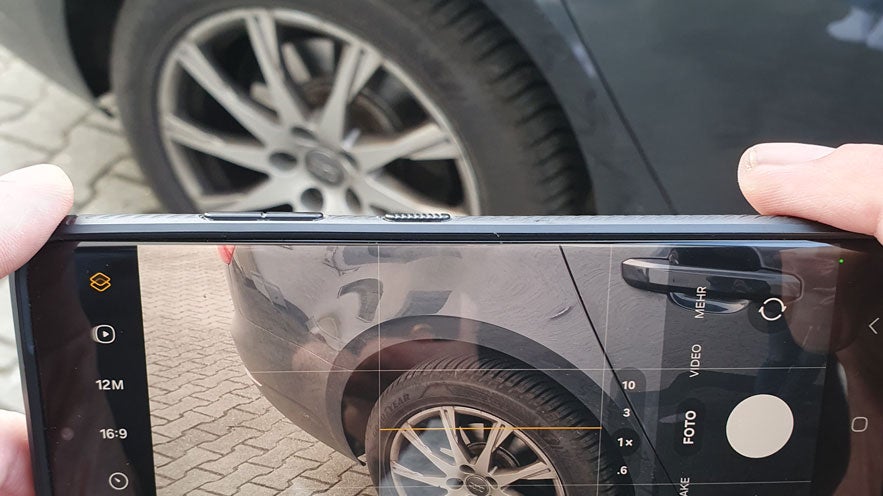 Smartphone fotografiert einen Unfallschaden an einem Autokotflügel mit sichtbaren Kratzern und Felge im Bildausschnitt.