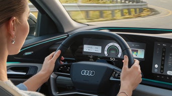 The cockpit of the Q6 SUV e-tron, viewed from the rear seat. A young woman is sitting in the driver's seat, holding the steering wheel with both hands. Through the windshield, you can see a road winding through the countryside.