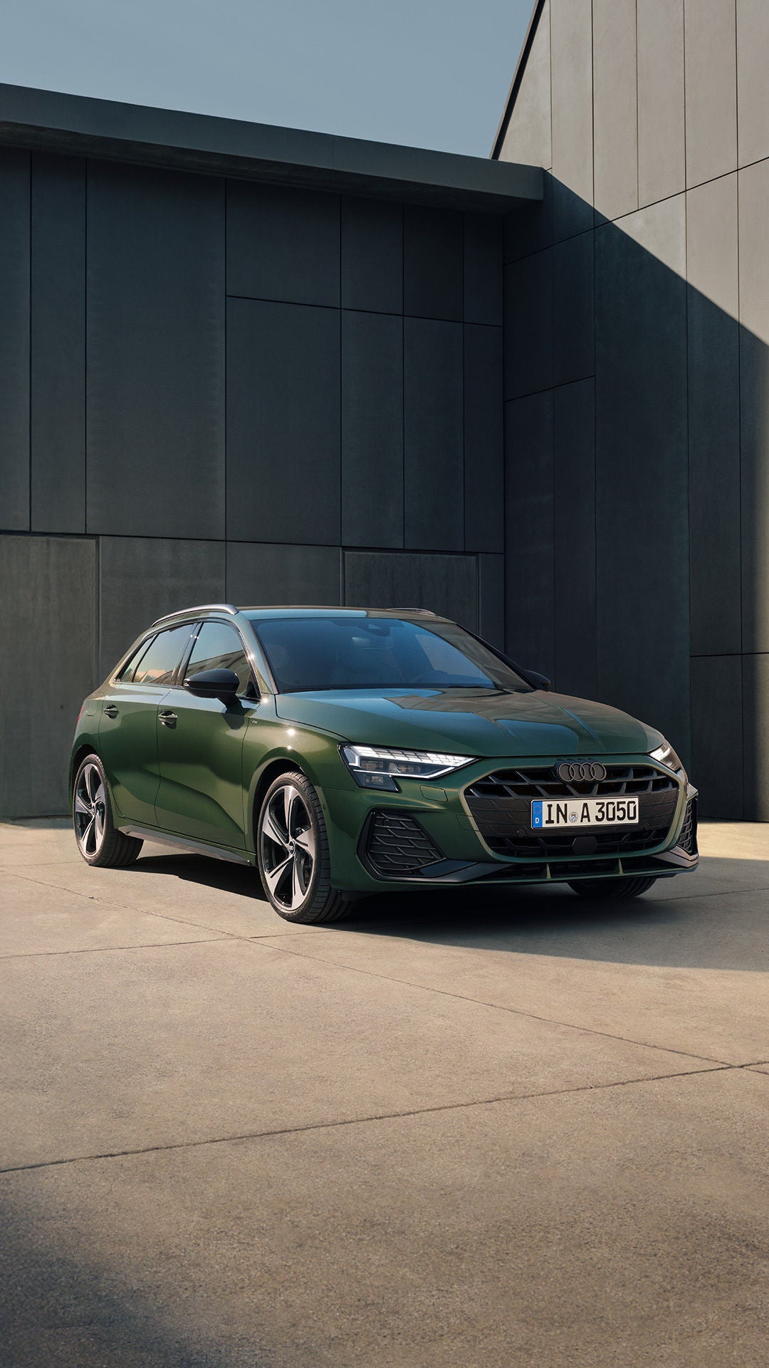 Grüner Audi A3 steht vor einer modernen, dunklen Gebäudewand auf hellem Betonboden bei Tageslicht.