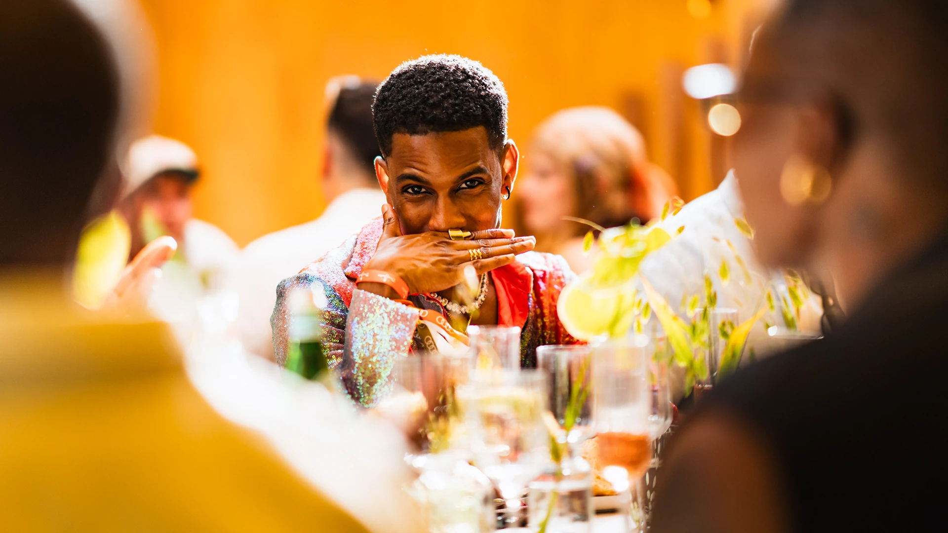 A woman in a tan suede jacket smiles over her shoulder at a dinner table decorated with bright orange and yellow flowers, surrounded by other guests. A woman in a tan suede jacket smiles over her shoulder at a dinner table decorated with bright orange and yellow flowers, surrounded by other guests.
