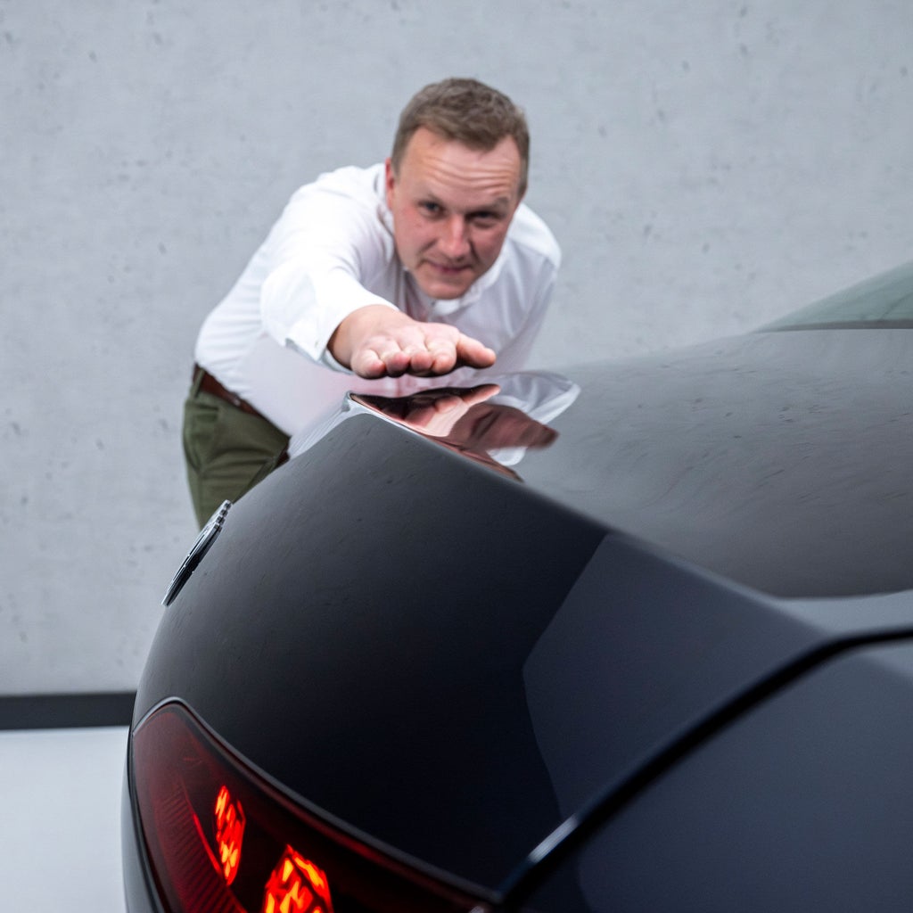 Sebastian Weiper steht neben einer Audi A6 Limousine und hält seine ausgestreckte Hand über das Heck des Fahrzeugs parallel zu dessen Form.