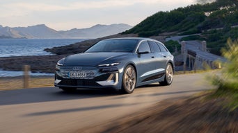 An Audi S6 Avant e-tron, sleek and modern, drives along a coastal road with mountains in the background, showcasing its elegant design and performance.