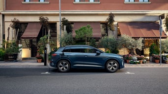 SUV Audi Q4 e-tron bleu en train de se recharger à une borne publique, garé dans une rue en milieu urbain devant un immeuble agrémenté de végétation et de vitrines. 