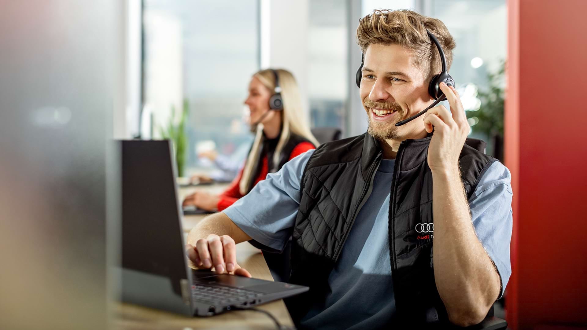 A blonde male person sitting in front of a laptop and talking via a wireless headset