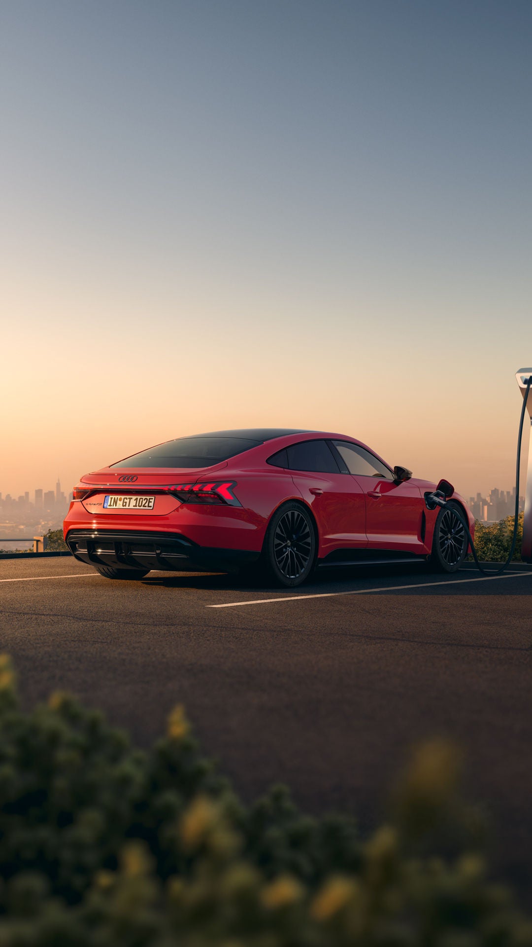 Ein roter Audi e-tron GT quattro steht auf einer Anhöhe über einer Stadt, die ins Licht eines Sonnenuntergangs gehüllt ist. Das Fahrzeug wird an einer öffentlicher Ladesäule geladen. Neben dem Fahrzeug stehen eine Frau und ein Mann.