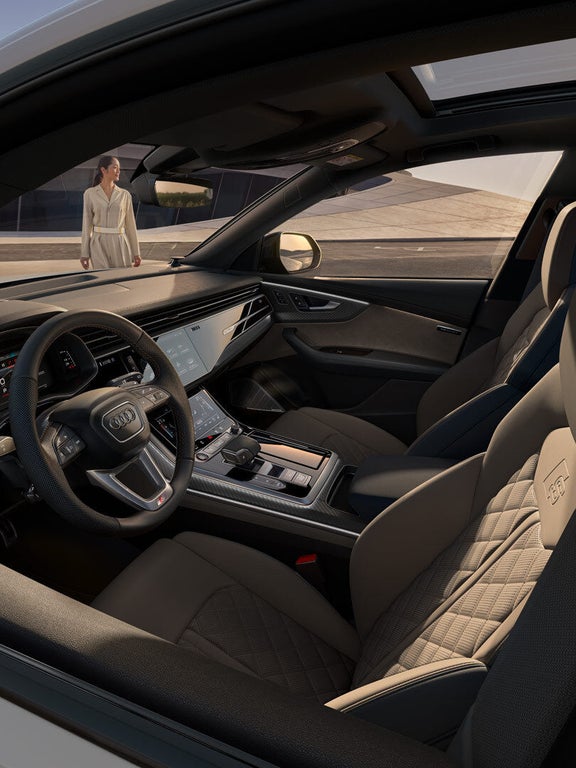 Innenraum eines Audi SQ8 SUV mit beigen Sportsitzen, großem Touchdisplay und digitalem Cockpit, Frau draußen im Hintergrund.