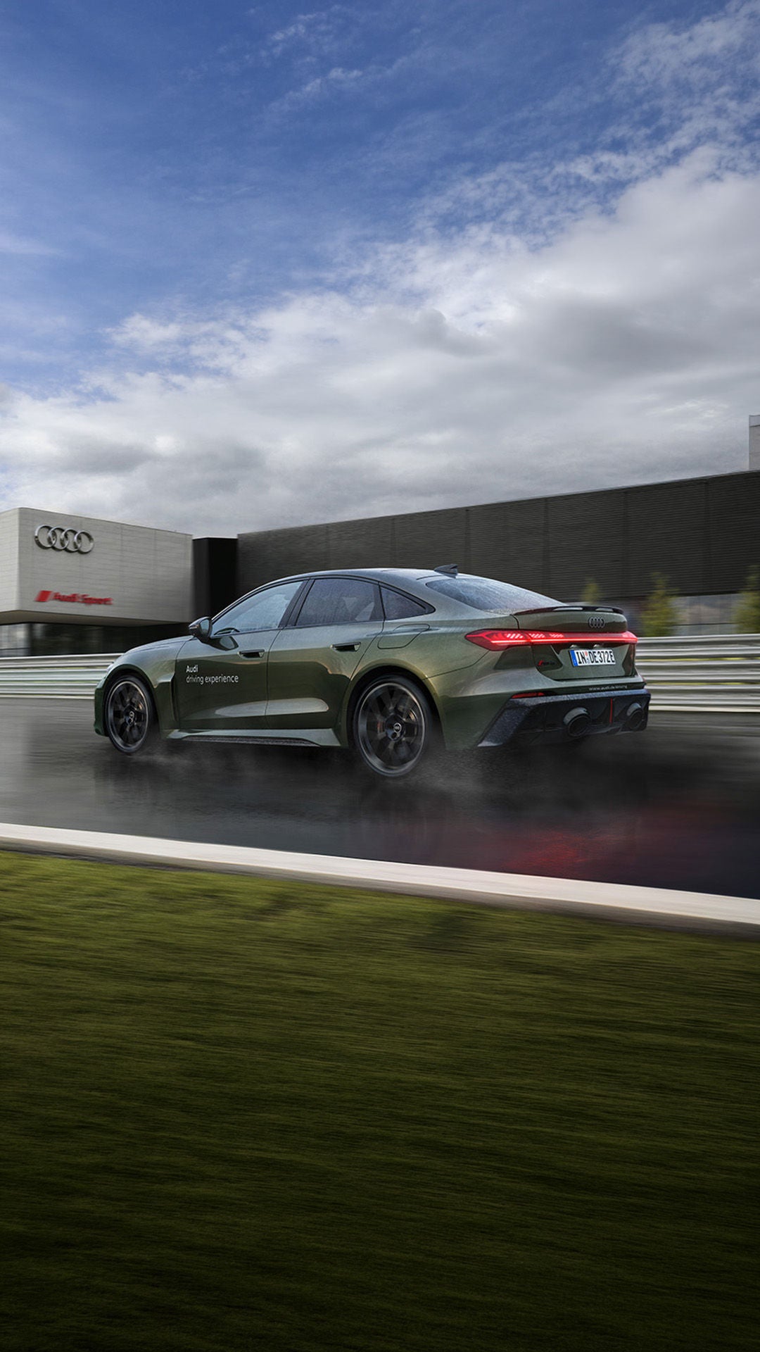 Dunkelgrüne Audi RS 5 Limousine fährt auf nasser Strecke vor Audi-Gebäude, Rücklichter leuchten.