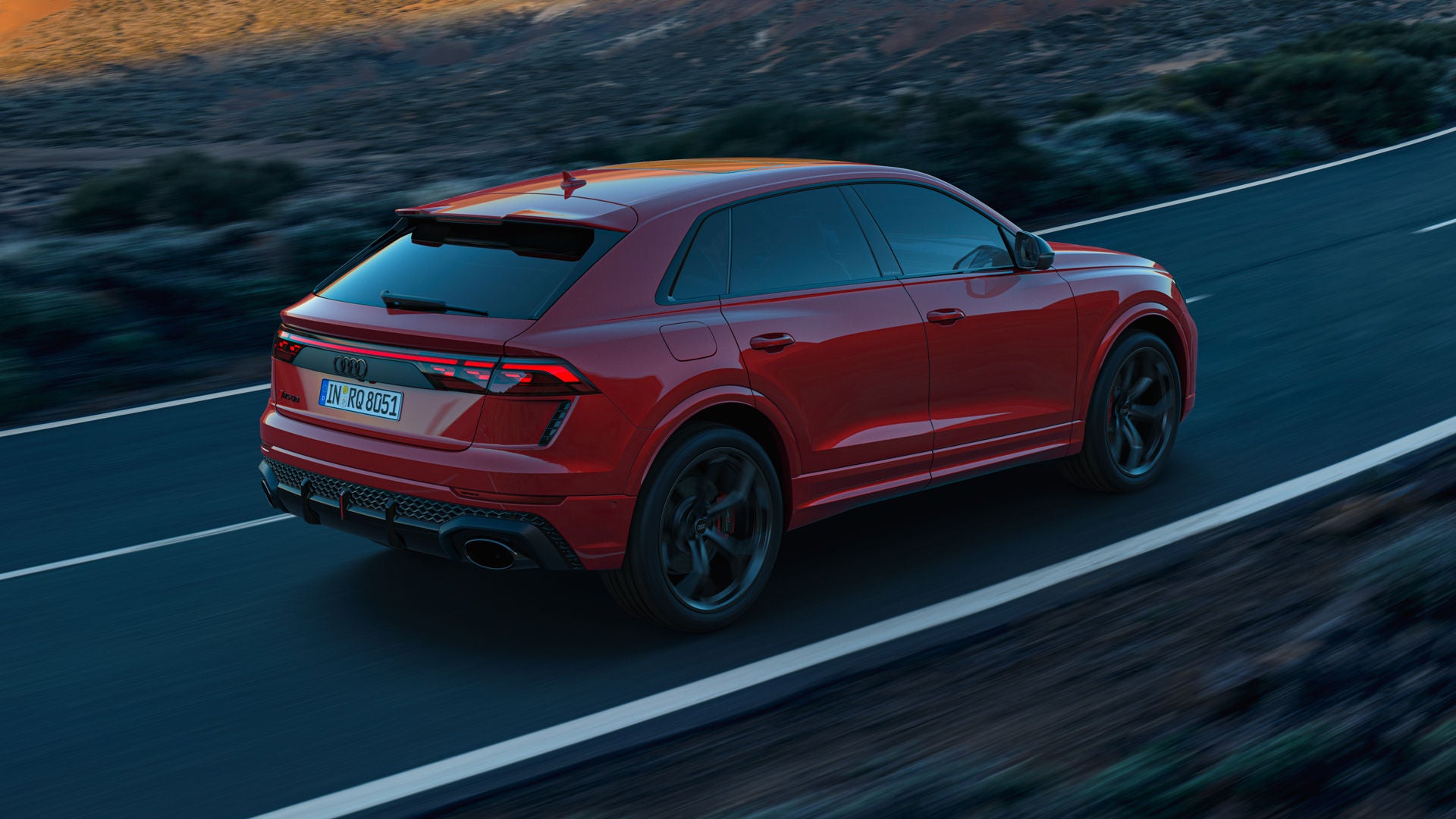 De RS Q8 SUV Performance, van achteren gezien vanuit een hoek op een bergweg.  De zon verlicht het rotsachtige landschap op de achtergrond, terwijl het voertuig in de schaduw blijft.