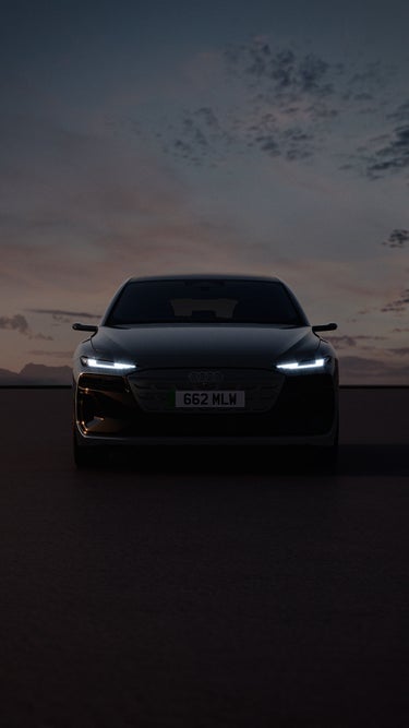 A black Audi A6 Avant e-tron car is illuminated against a dusky sky, showcasing its modern design and LED headlights.