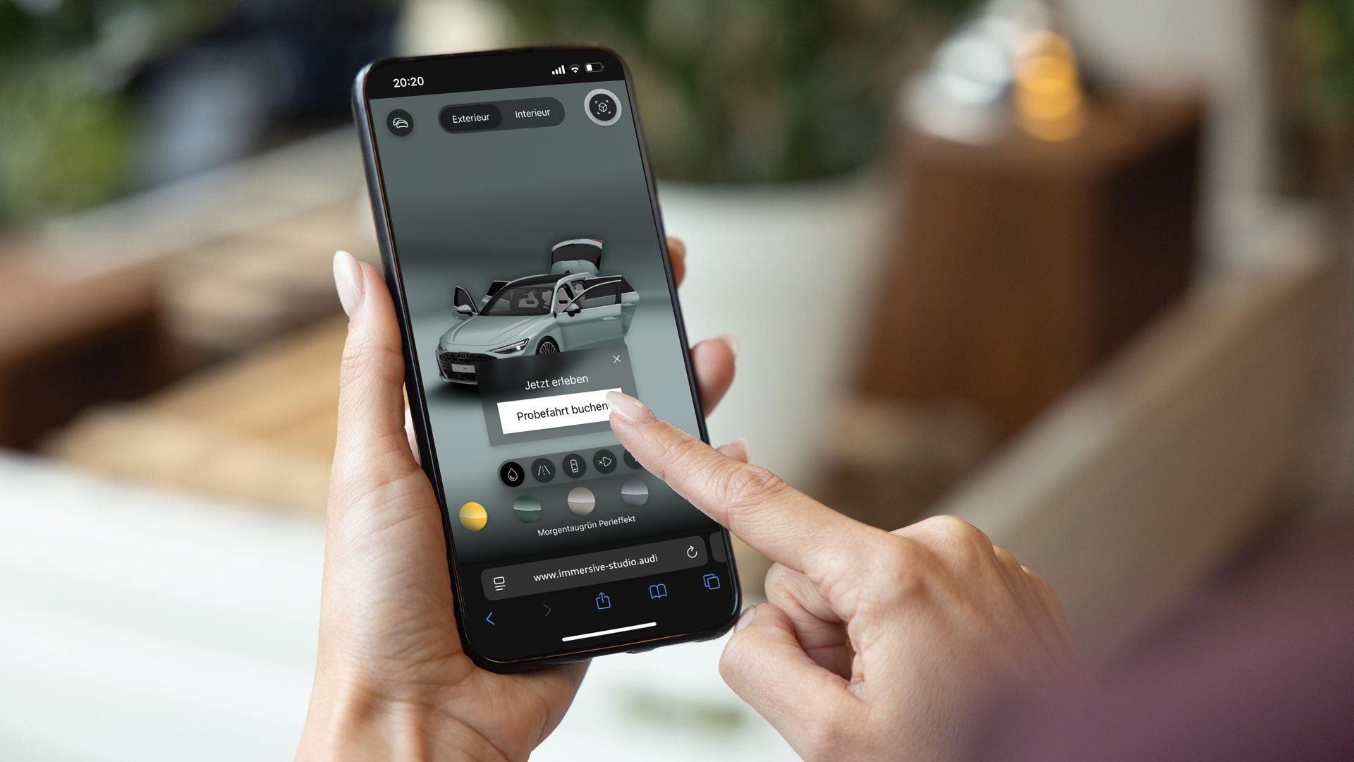 Eine Person hält ein Smartphone in der Hand und nutzt eine App zur virtuellen Autokonfiguration. Auf dem Bildschirm ist ein 3D-Modell eines silbergrauen Autos mit geöffneten Türen, Kofferraum und Motorhaube zu sehen. Die App bietet verschiedene Auswahlmöglichkeiten für Farben und Funktionen, und die Person tippt auf die Schaltfläche „Probefahrt buchen“. Oben im Display stehen die Optionen „Exterieur“ und „Interieur“. Die Website „immersive-studio.audi“ ist am unteren Bildschirmrand sichtbar.