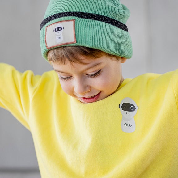 Nahaufnahme eines Kindes, mit ausgebreiteten Armen, das einen gelben ADUI Kids Pullover und grünen Beanie trägt.