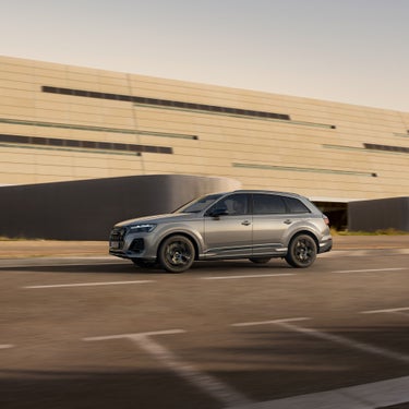 Ein silberner Audi Q7 SUV TFSI e fährt auf leerer Straße, modernes Gebäude im Hintergrund, weiches Abendlicht.