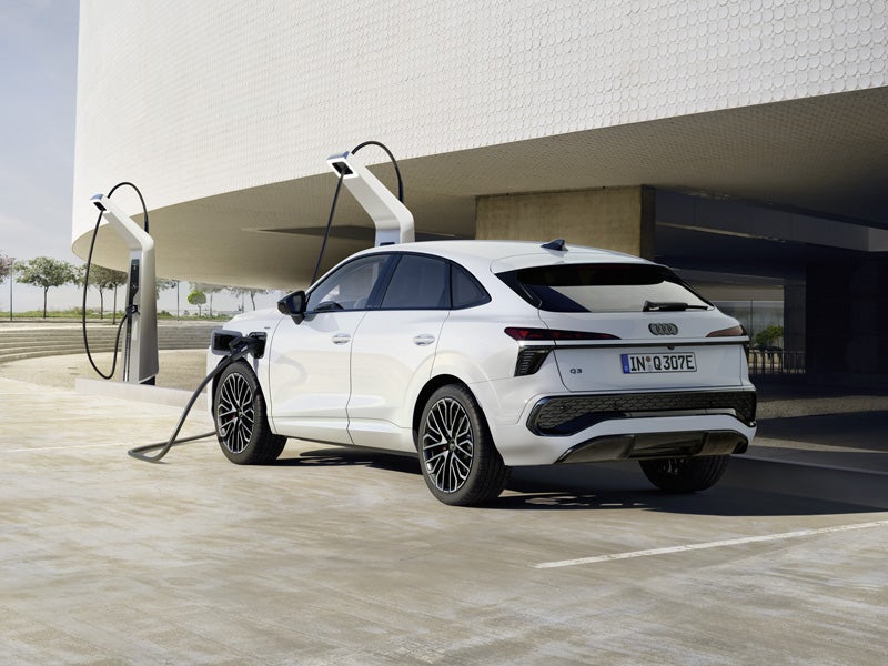Una Q3 Sportback e-hybrid bianca sta ricaricando in una colonnina di ricarica di fronte a un edificio moderno, il veicolo proietta lunghe ombre.
