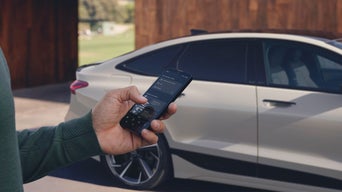 Person hält Smartphone mit Fahrzeug-App in der Hand, im Hintergrund ein geparkter weißer Audi.