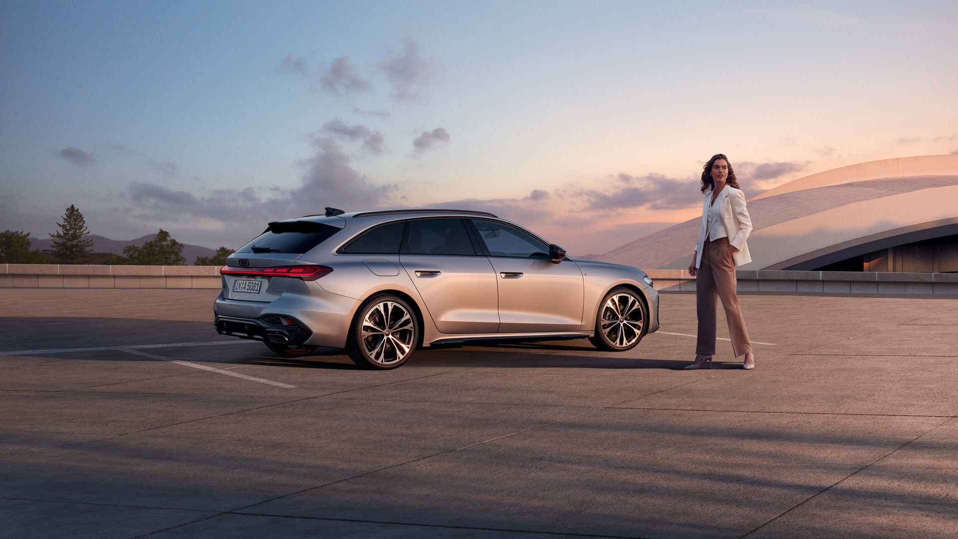 Eine Frau steht neben einem silbernen Audi Modell bei Sonnenuntergang und guckt in die Ferne