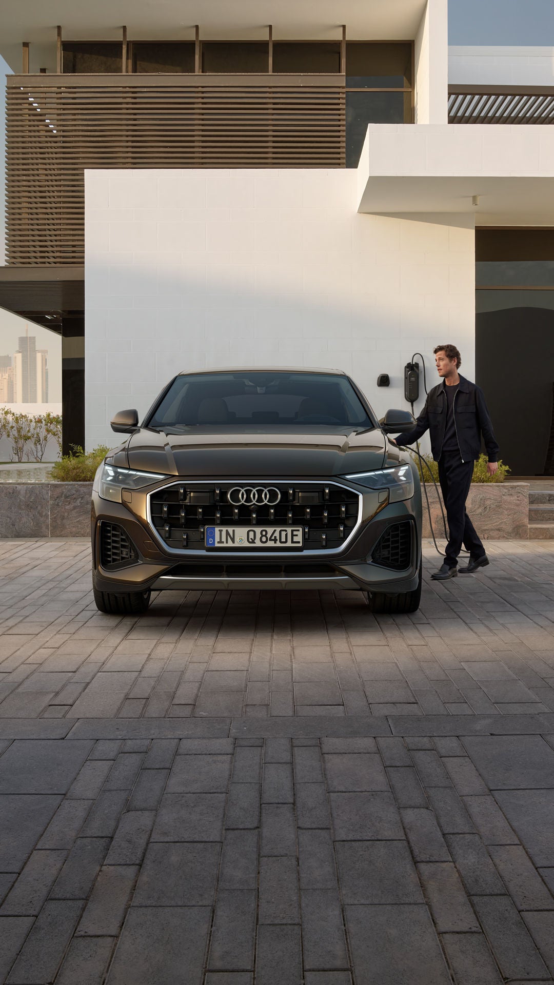 Vooraanzicht van de Q8 SUV e-hybride, met daarachter een modern woongebouw.  Naast het voertuig staat een modieus geklede jongeman die de oplaadstekker vasthoudt van een laadstation dat op het gebouw is gemonteerd.  Op de achtergrond aan de zijkant bevindt zich een kleine tuin en daarachter is de skyline van een grote stad te zien.
