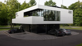 Ein weisses kubushaftes Gebäude mit einer verspiegelten Glasfläche. Danben sind auf der Wand die 4 Audi Ringe angebracht. Audi charging hub Nürnberg