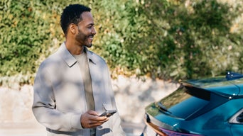 Ein Mann steht mit einem Handy in der Hand neben einem blauen Audi, im Hintergrund eine grüne Hecke.