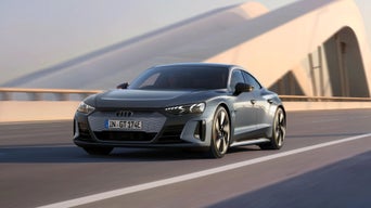 A silver Audi e-tron GT vehicle driving on a sunlit road with modern architecture in the background