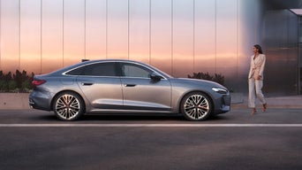 Eine Frau im beigen Anzug steht vor einem beigen Audi und schaut nach links