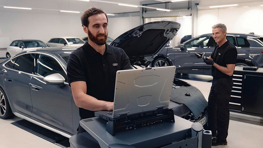 Mitarbeiter arbeitet am Laptop vor Audi, Kollege prüft Fahrzeug im Hintergrund.