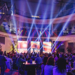 Two women raise their arms and stand on a stage with colorful lighting effects in front of an audience at the Audi Forum Neckarsulm.