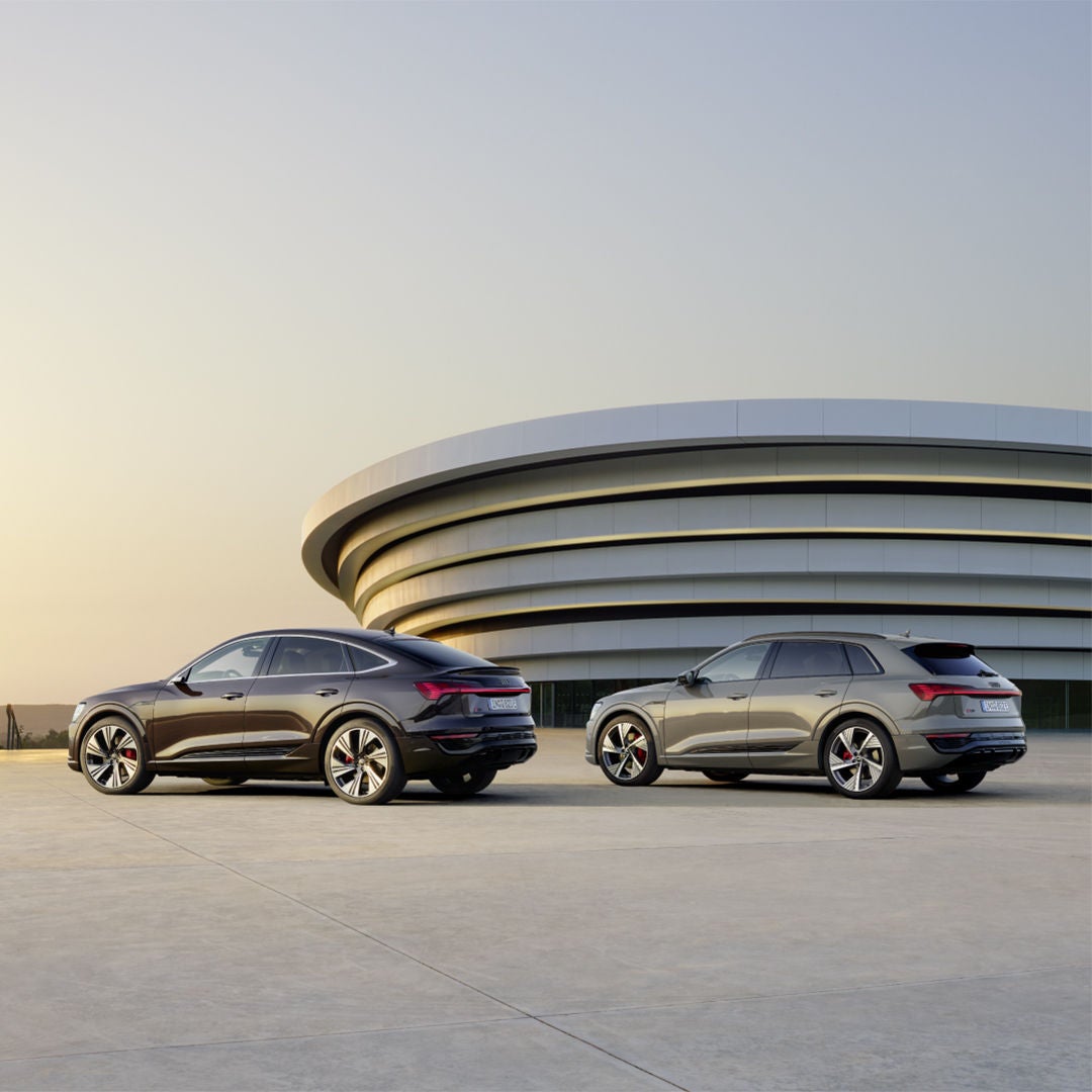 Zwei Audi Q8 e-tron stehen nebeneinander auf einer Fläche vor einem modernen, runden Gebäude bei Sonnenuntergang.