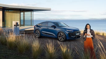 Frau mit Smartphone vor einem blauen Audi Q6 SUV e-tron, geparkt an Ladestation neben modernem Haus mit Meerblick.