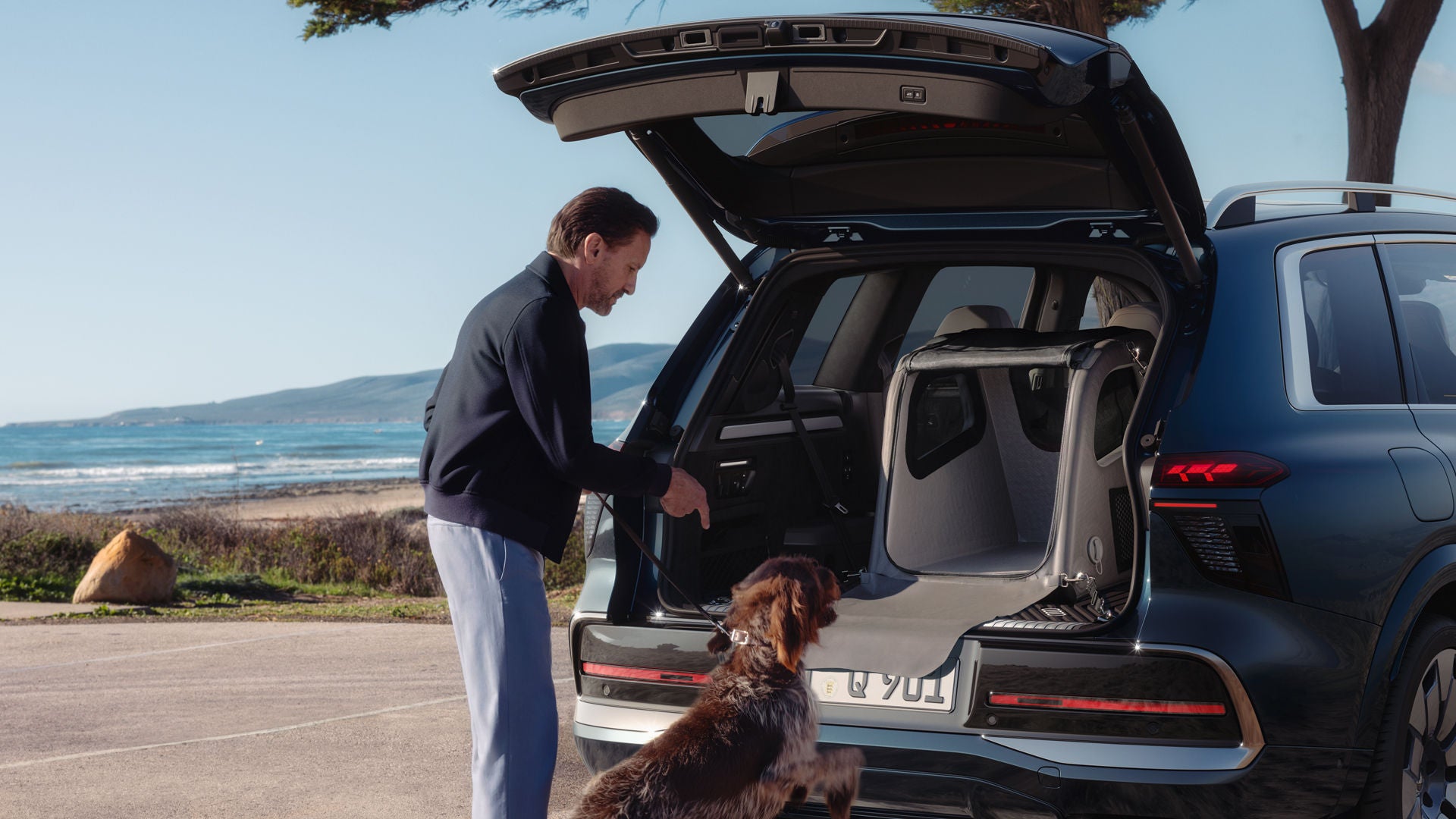 Un homme se tient debout devant le coffre ouvert du SUV Audi Q9. Son chien s'apprête à sauter dans une cage pour chien placée dans le coffre.
