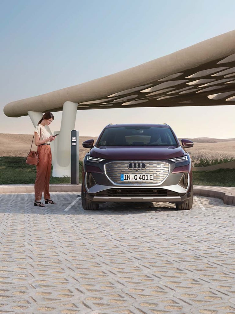 Frau steht mit Smartphone neben ladendem, violetten Audi Q4 e-tron vor Ladesäule in wüstenartiger Umgebung mit moderner Überdachung.