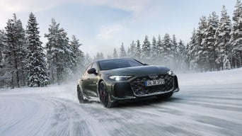 A dark green Audi RS 5 sedan drives along a snow-covered road; snow dust, a pine forest, and a blue sky.