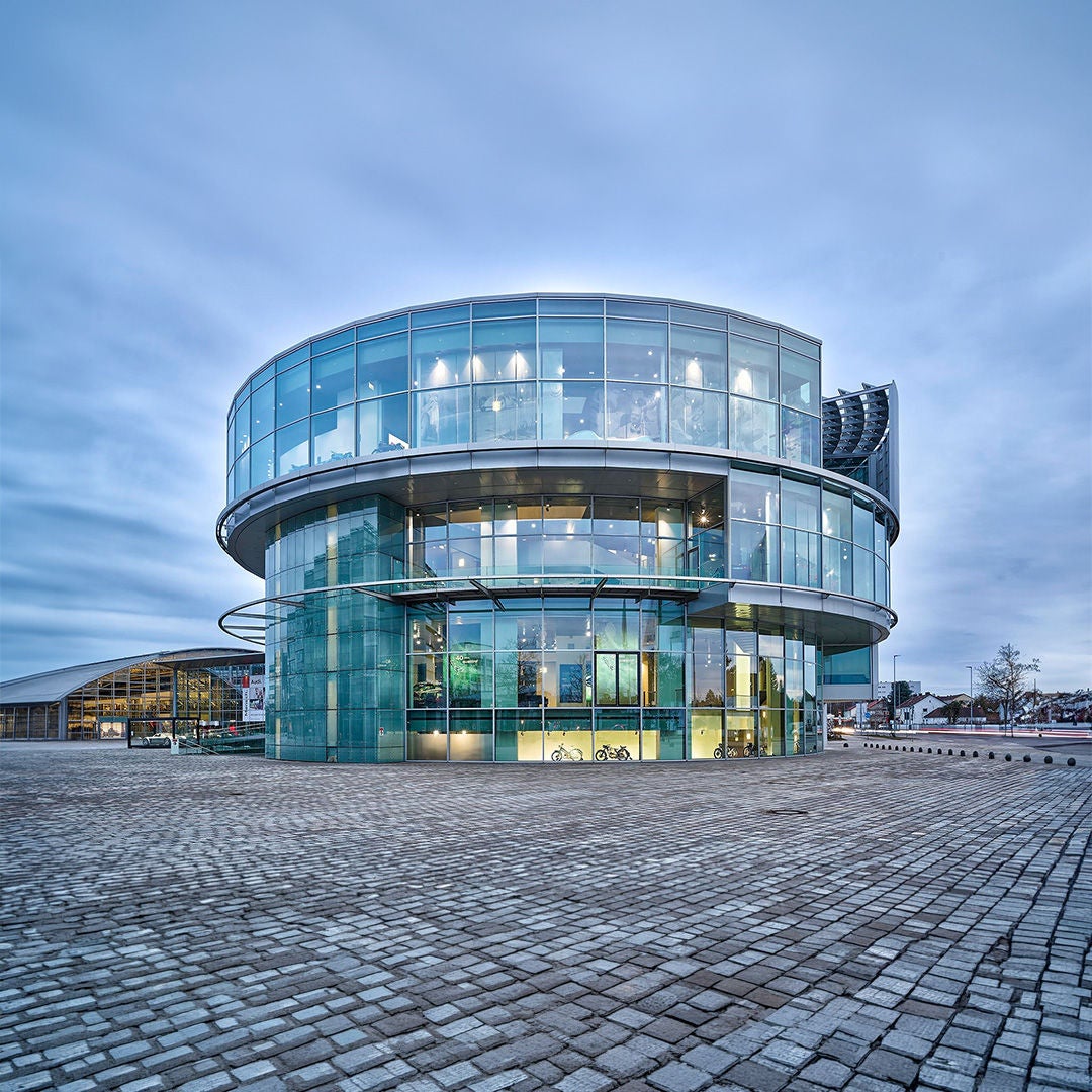 Rundes modernes Glasgebäude auf gepflastertem Platz bei bewölktem Himmel.