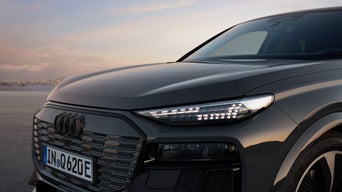 Close-up of a sleek grey Audi Q6 Sportback e-tron with a distinct grille and modern LED headlights, set against a soft sunset backdrop.