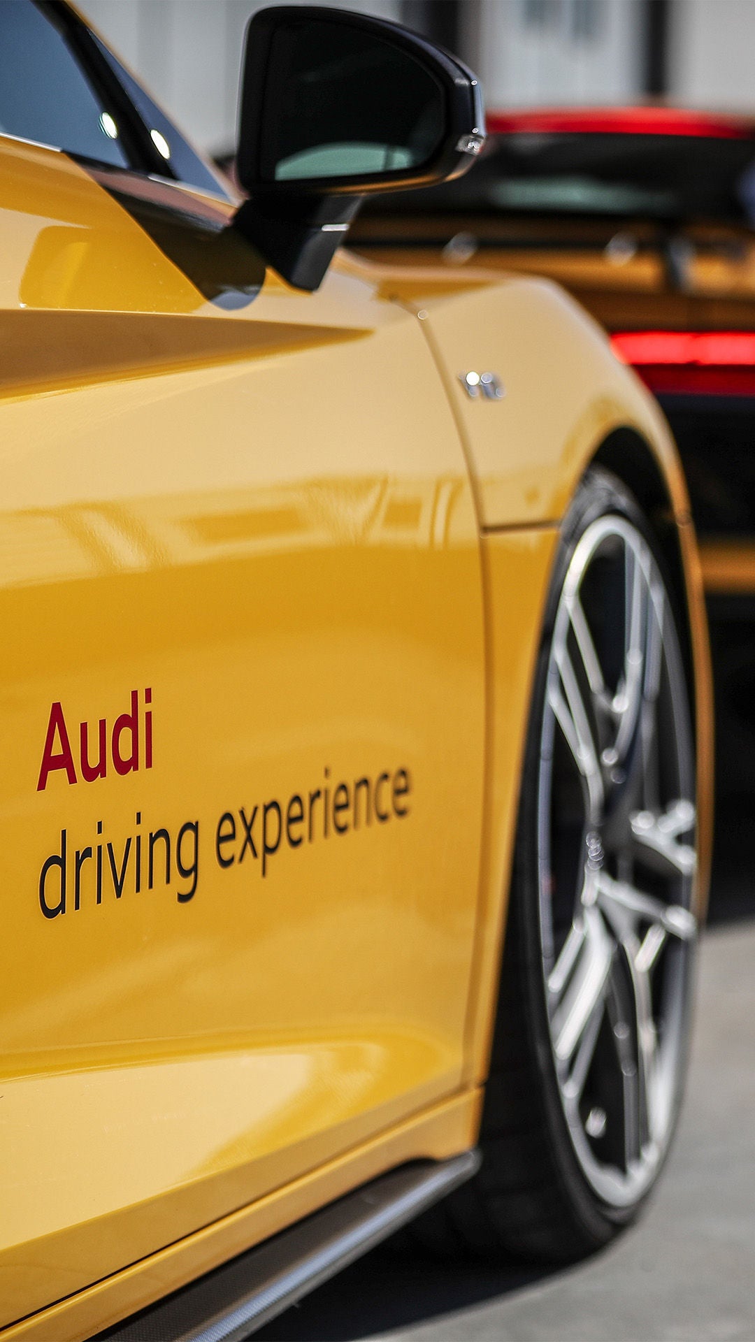 Gelber Audi Sportwagen mit Schriftzug „Audi driving experience“, Nahaufnahme; weitere gelbe Fahrzeuge in Reihe im Hintergrund.