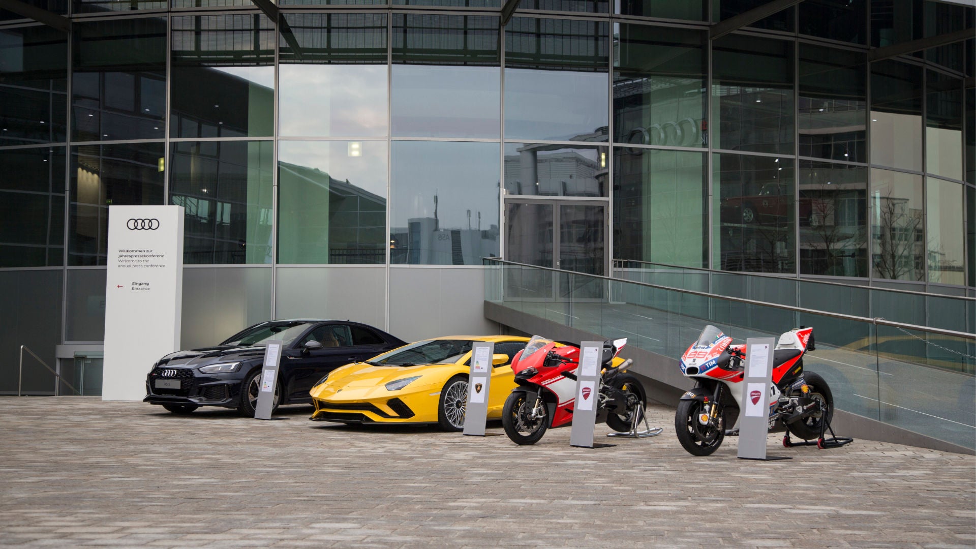  An Audi, a Lamborghini and two Ducati motorbikes in front of the entrance to the Audi Forum Ingolstadt
