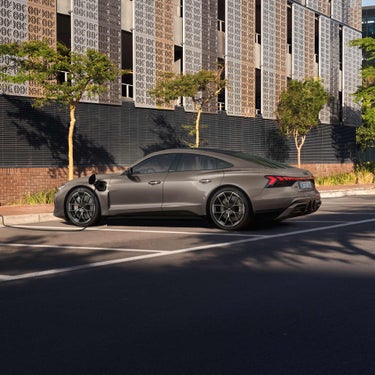 Ein lila Audi RS e-tron GT lädt an einer Straßenladestation vor einer modernen Metallfassade mit Bäumen.