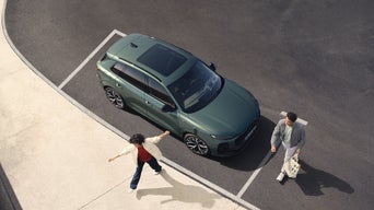 Vus du dessus, deux personnes se tiennent sur un parking. Une femme, les bras tendus, se tourne vers un homme qui marche, un sac à la main. Une Audi Q3 vert foncé est garée à côté d'eux.