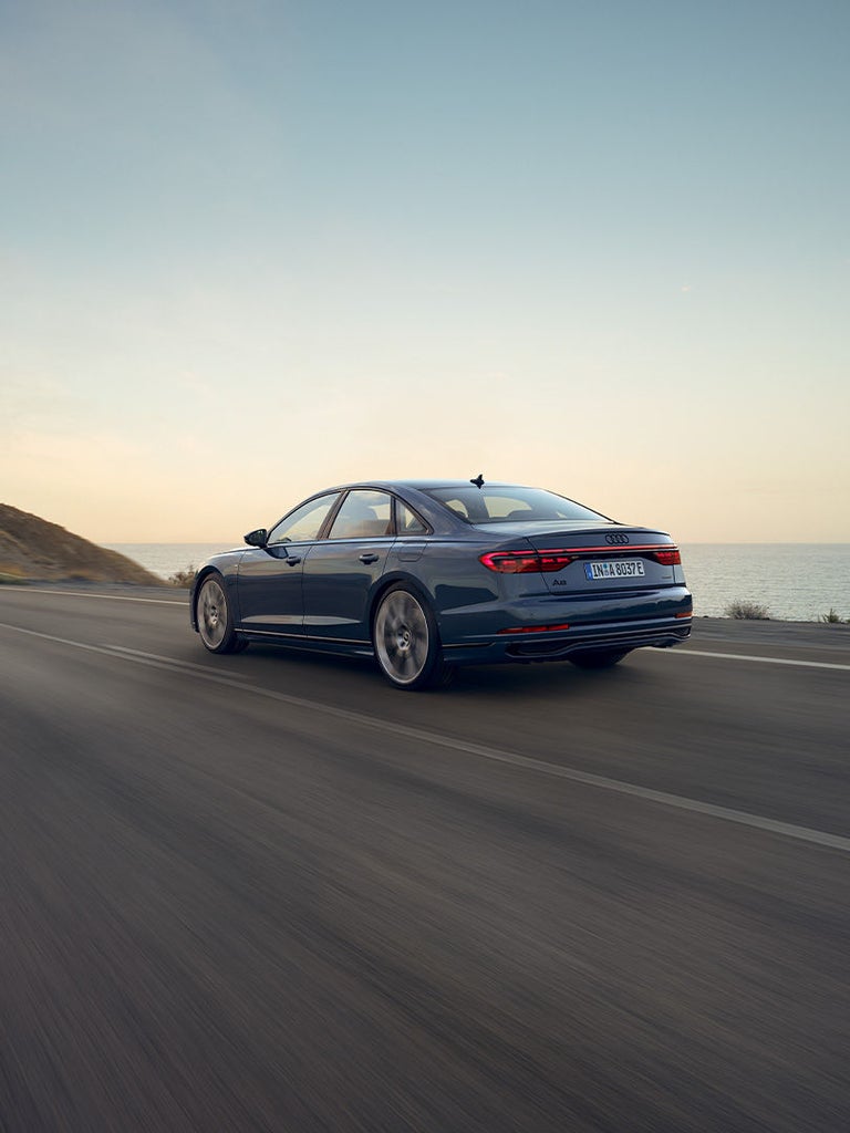Ein dunkelblauer Audi A8 60 TFSI e fährt bei Sonnenuntergang auf einer Straße am Meer entlang.