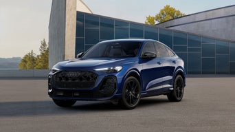 El Q5 Sportback, en la versión S line, se encuentra situado en ángulo sobre una superficie de asfalto frente a un gran edificio.