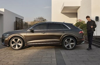 A brown Audi parked in a modern driveway, next to a contemporary house, surrounded by greenery.