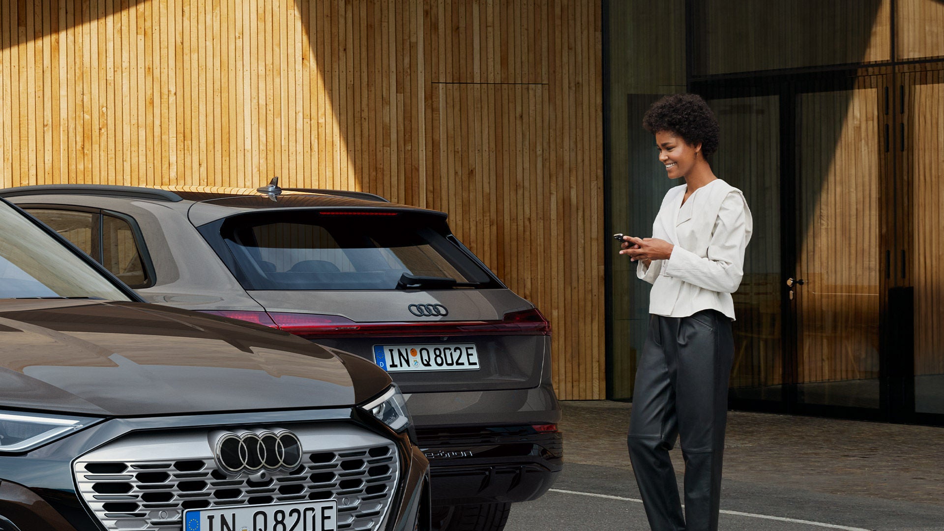 Frau steht lächelnd vor zwei geparkten Audi-Fahrzeugen und bedient ein Smartphone, moderner Holzbau im Hintergrund. 