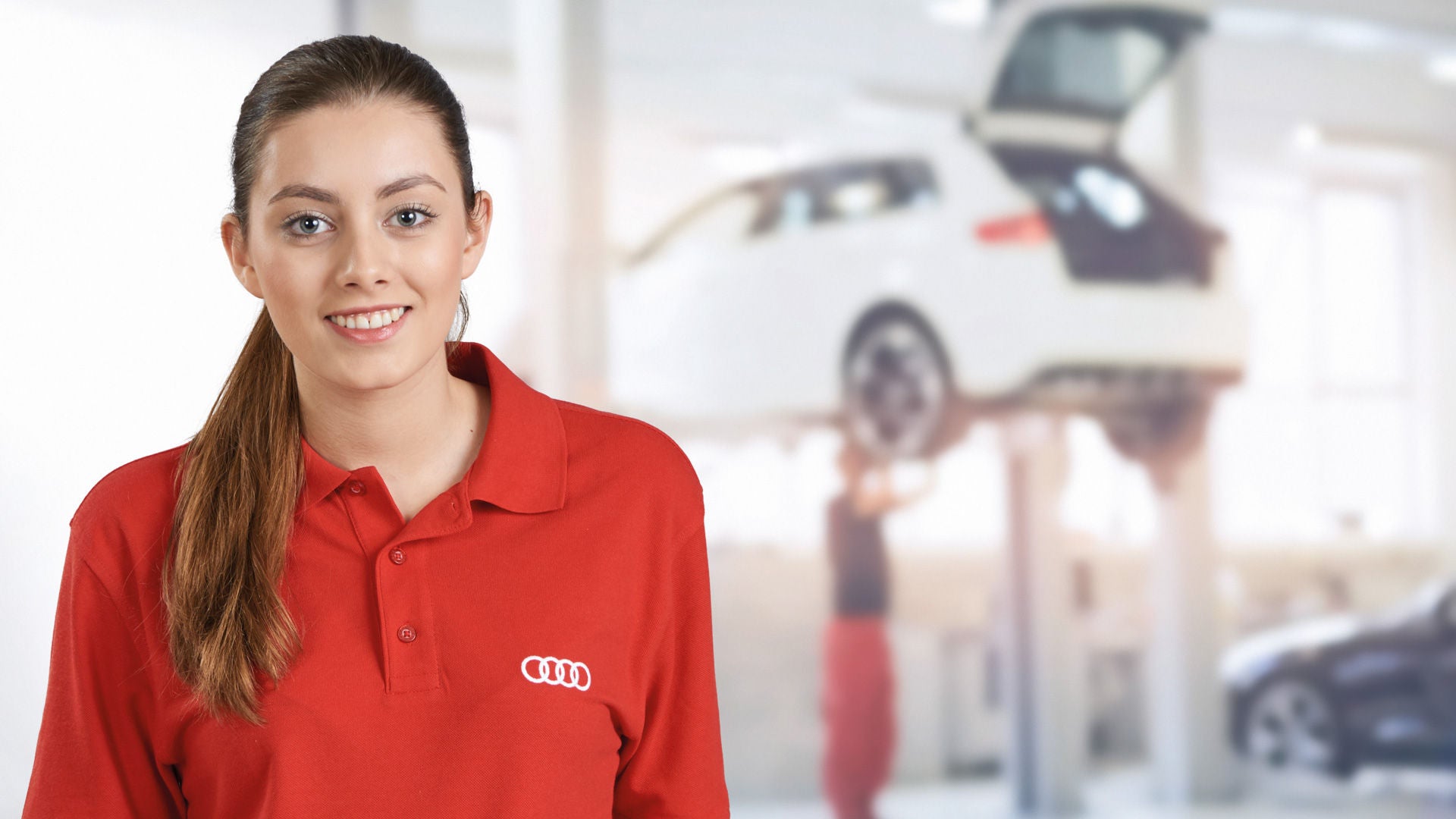 Frau schaut in die Kamera und lächelt. Im Hintergrund arbeitet ein KFZ Mechatroniker an einem Auto