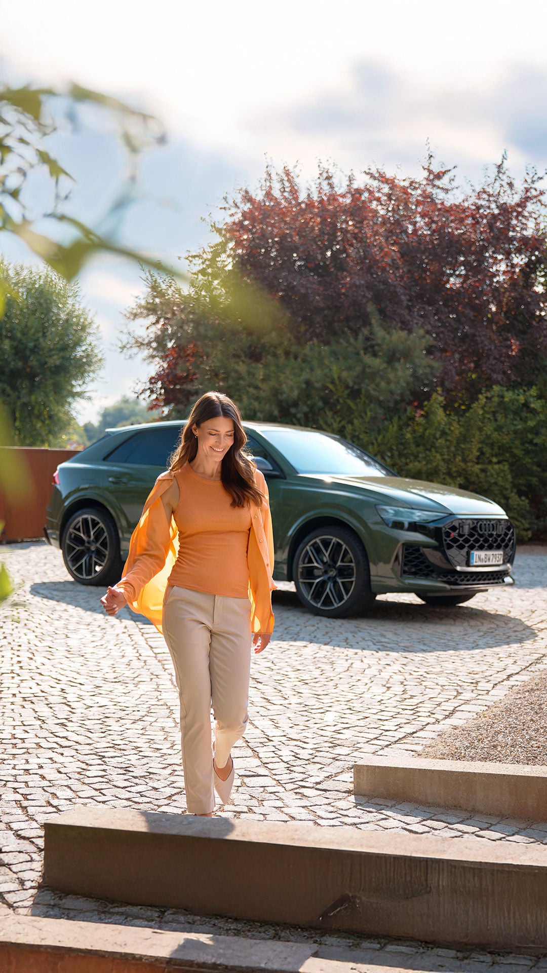 Eine Frau mit langen dunklen Haaren geht lächelnd über einen gepflasterten Hof, während im Hintergrund ein modernes Auto vor einem Haus geparkt ist.