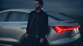 Mann lehnt abends an einem silbernen Audi mit leuchtendem Rücklicht, im Hintergrund hügelige Landschaft.