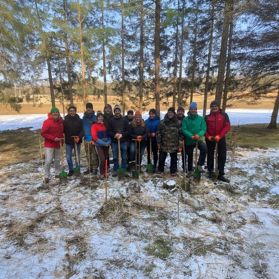 Gruppe von 13 Personen mit Spaten steht im verschneiten Wald für eine Baumpflanzaktion, winterlich gekleidet und lächelnd.