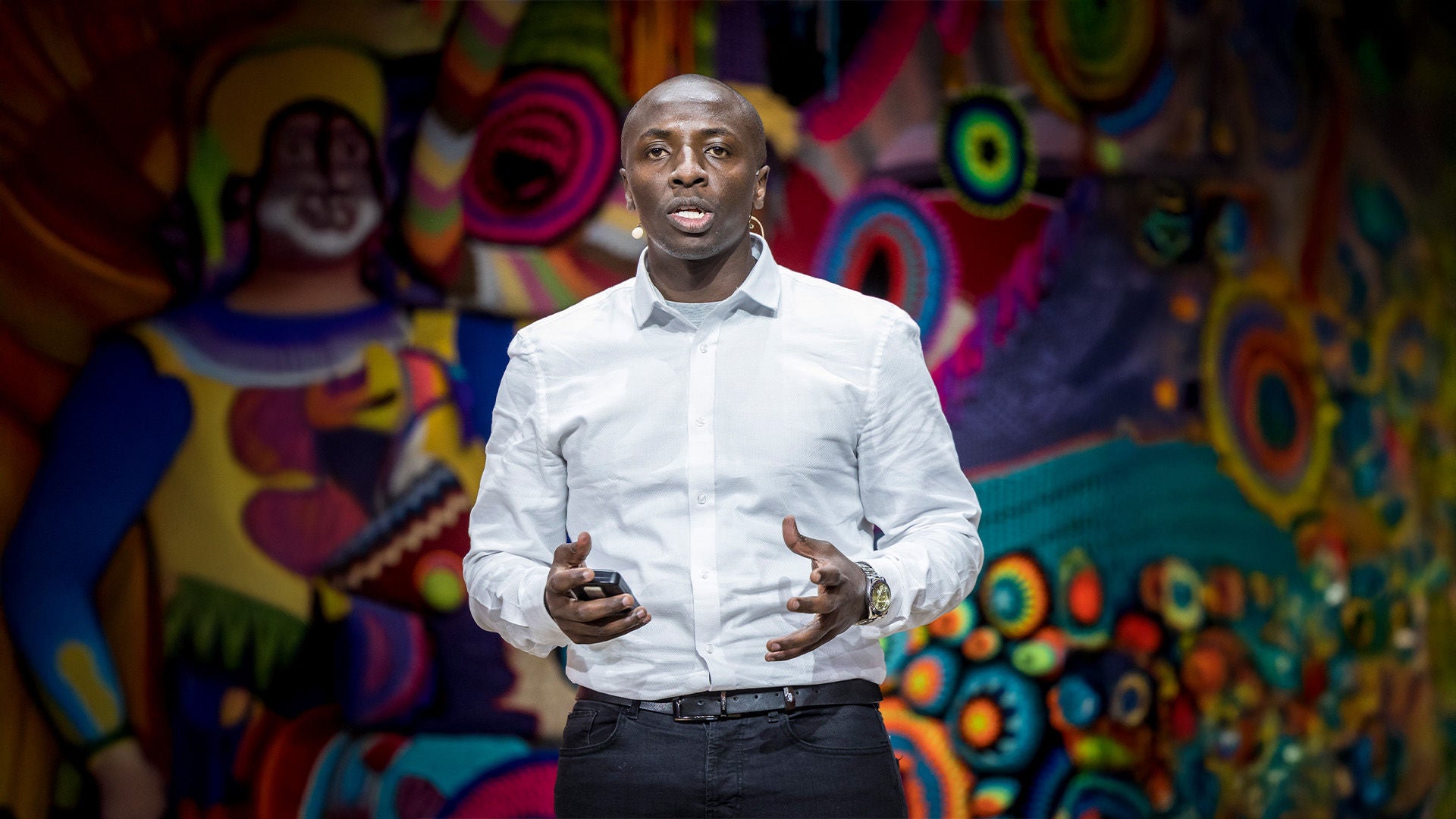 Mann im weißen Hemd spricht auf einer Bühne vor farbenfrohem, abstraktem Hintergrund.